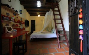 Un dormitorio con una cama bajo un dosel blanco, una escalera de madera, un escritorio rojo con una silla y una estantería con diversos objetos.