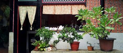Fachada de ladrillo con un pequeño porche. Hay plantas en macetas en el porche, una ventana con cortina y un celosía decorativa sobre la ventana.