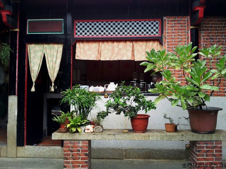 Fachada de ladrillo con un pequeño porche. Hay plantas en macetas en el porche, una ventana con cortina y un celosía decorativa sobre la ventana.