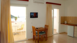 A room with a dining table, chairs, a TV, a kitchen area with a coffee maker, and a sliding glass door leading to a balcony with a view of the sea.