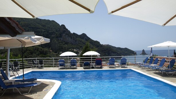 A pool area with blue lounge chairs and white umbrellas, overlooking a mountainous landscape and the sea.