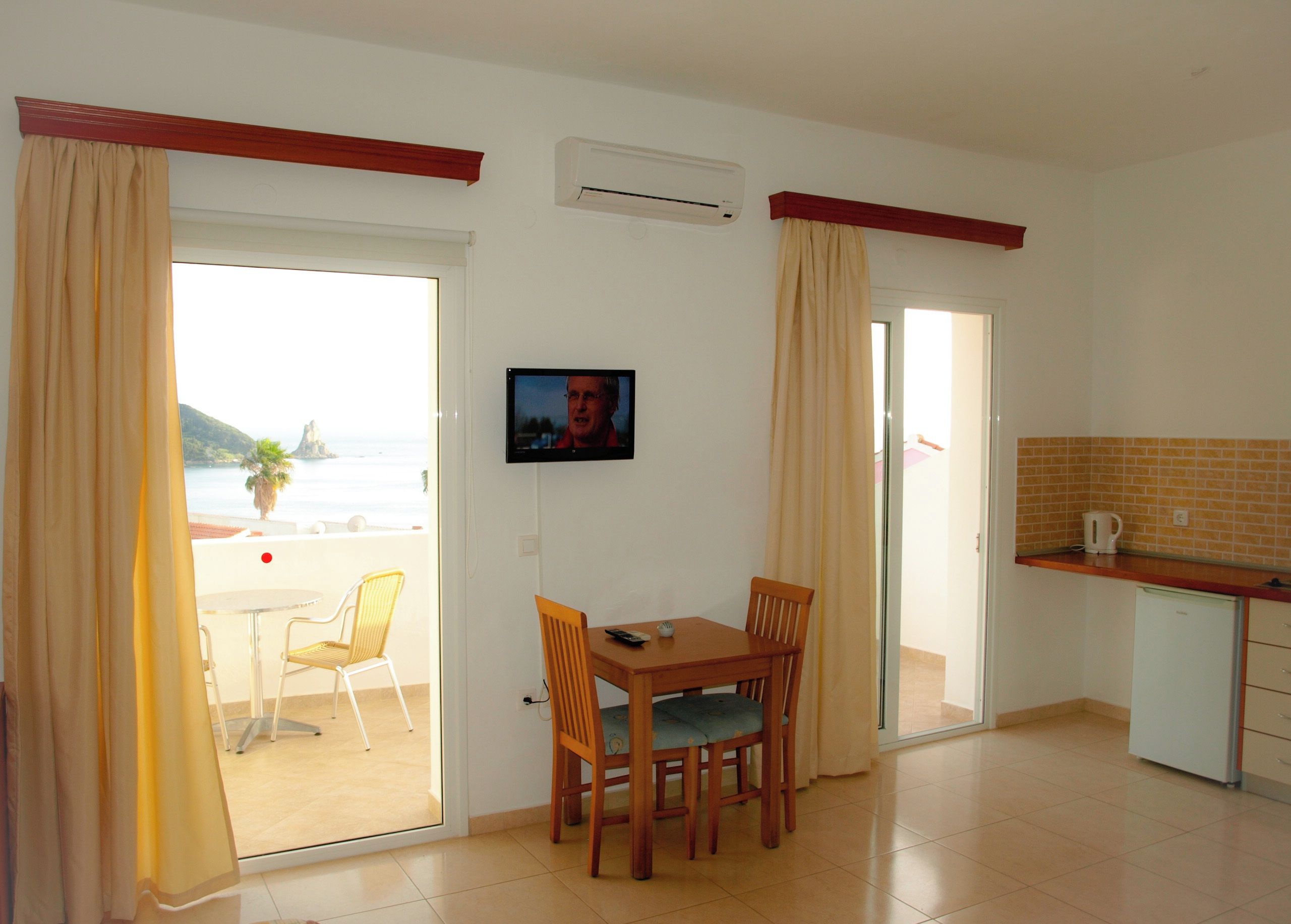 Una habitación con mesa de comedor, sillas, televisión, una zona de cocina con cafetera y un ventanal corredero que da a un balcón con vistas al mar.