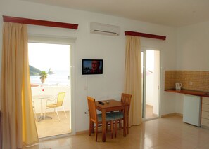 A room with a dining table, chairs, a TV, a kitchen area with a coffee maker, and a sliding glass door leading to a balcony with a view of the sea.