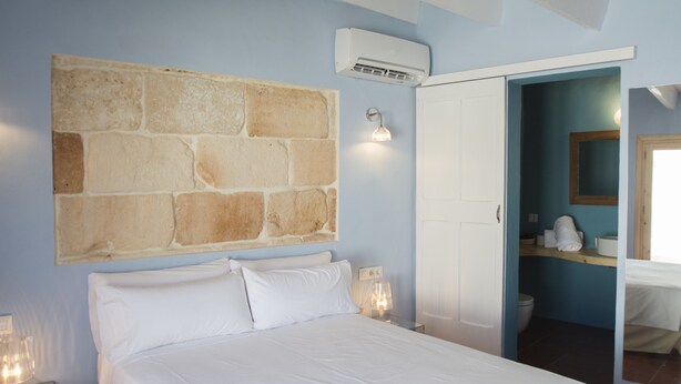 A bedroom with a large stone wall, a bed, a nightstand with a lamp, and an open door leading to a bathroom. - Rifugio Azul (Ciutadella de Menorca)