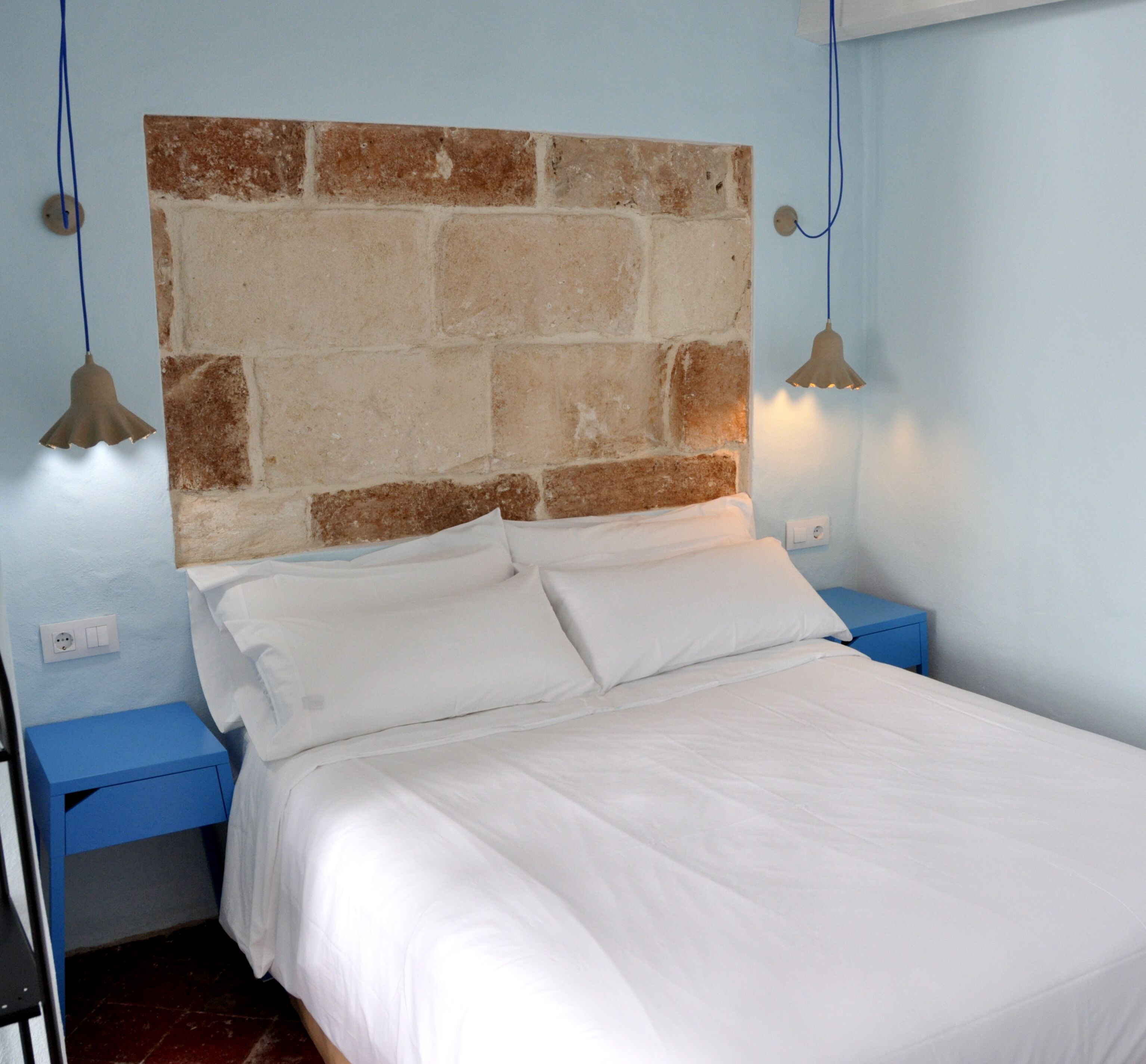 Un lit avec du linge de lit blanc, une tête de lit en pierre et deux tables de chevet avec des lampes.