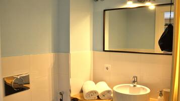A bathroom with a white sink, a mirror, and two rolled towels on a wooden shelf.