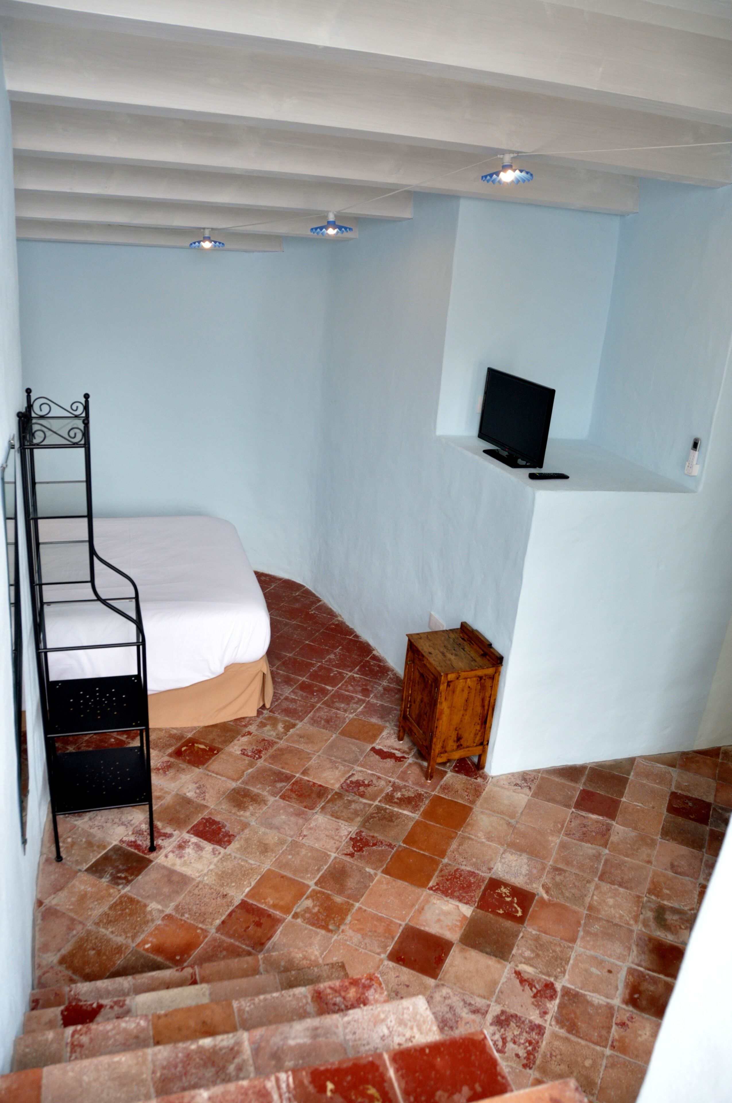 A small, narrow room with a bed, a wooden stool, and a television on a white stand.