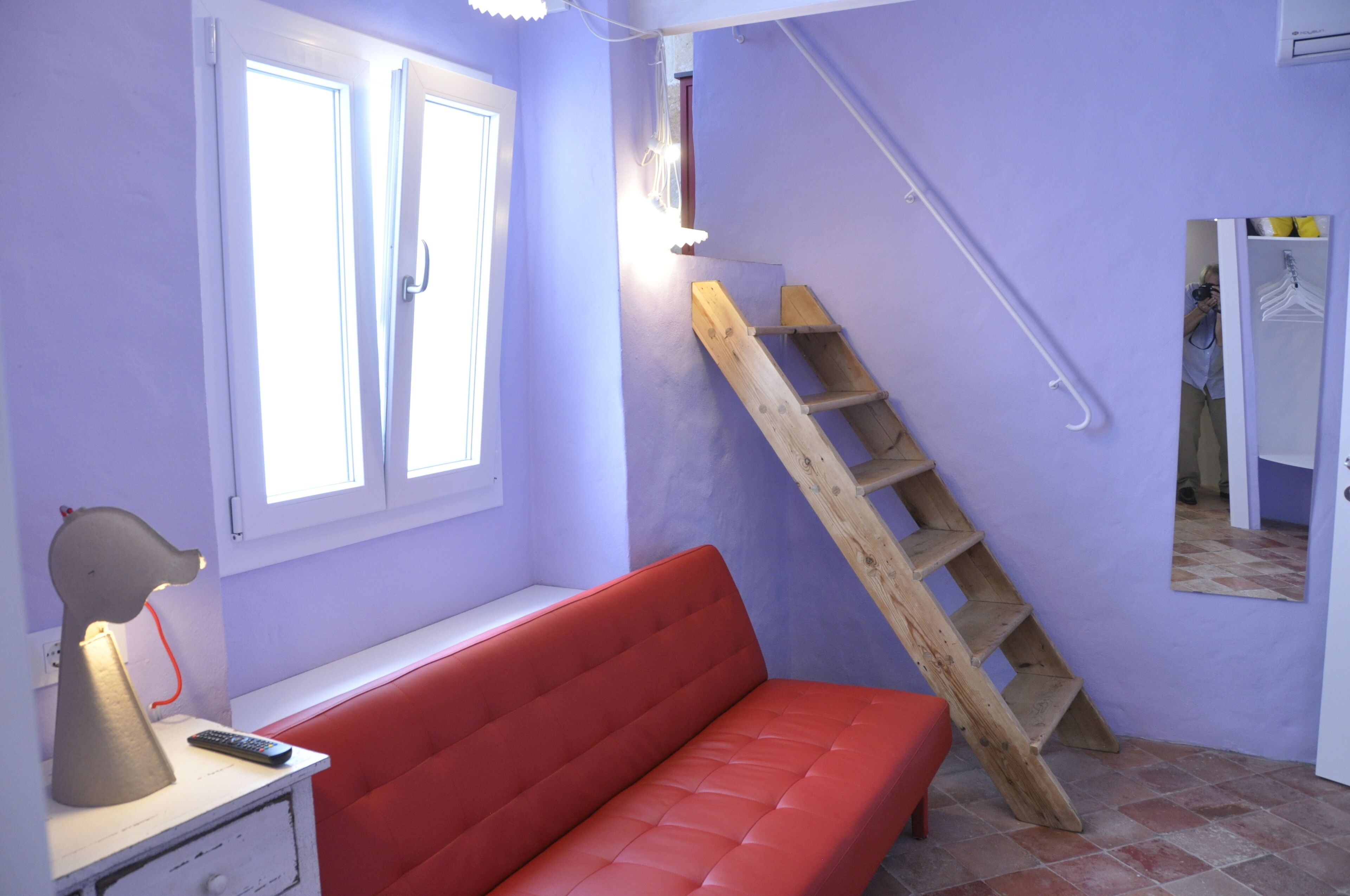 Una habitación con un sofá rojo, una escalera de madera, un ventanal con contraventanas blancas, una lámpara y un espejo.