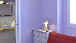 A room with a red sofa, a white bedside table with a lamp, a mirror above a sink, and a window with natural light. - Rifugio Azul (Ciutadella de Menorca)