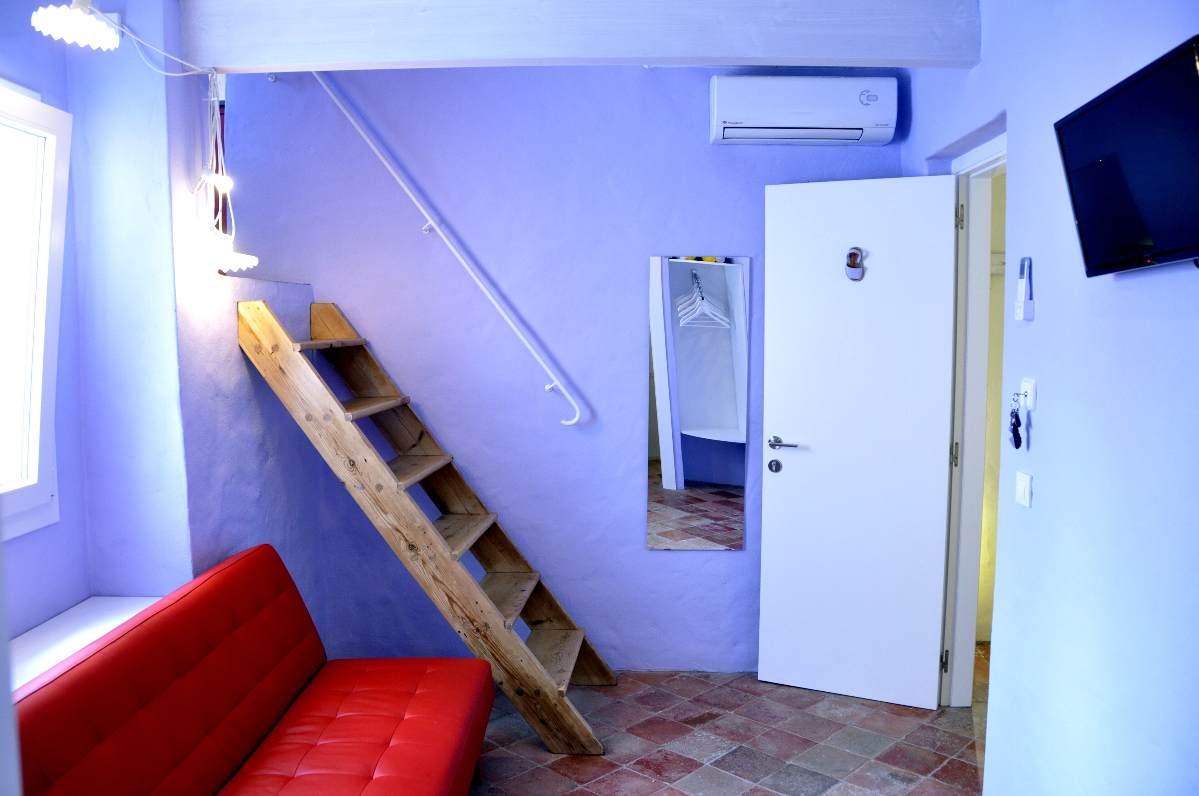 Quarto compacto com um banco vermelho, escada de madeira, porta branca e TV fixada na parede.
