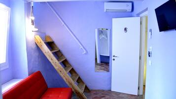 Quarto compacto com um banco vermelho, escada de madeira, porta branca e TV fixada na parede.