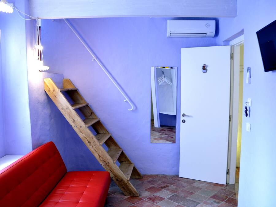 Quarto compacto com um banco vermelho, escada de madeira, porta branca e TV fixada na parede.