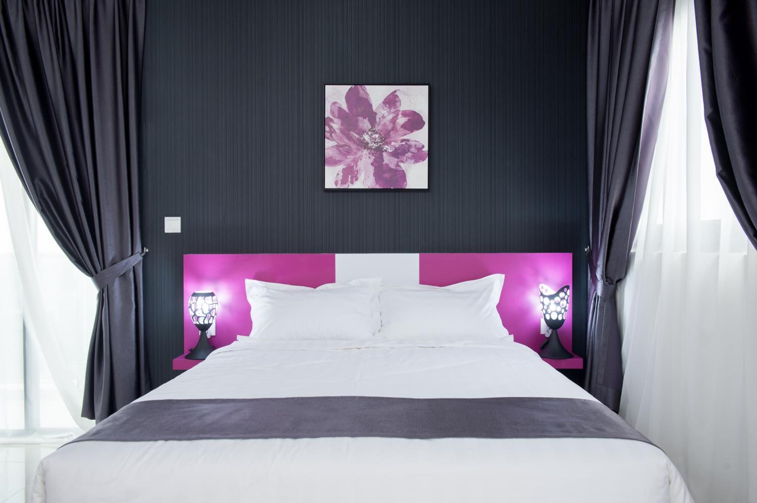 A modern hotel room with a large bed, dark walls, and a pink accent wall.