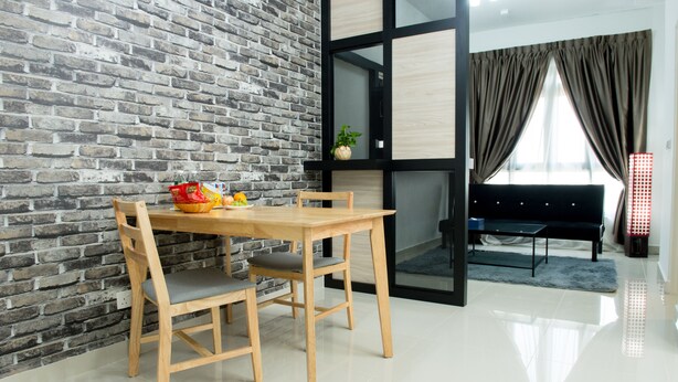A dining area with a brick wall, a wooden table with chairs, and a view of a living room with a sofa and curtains. - Ezzyhome Medini (Iskandar Puteri)