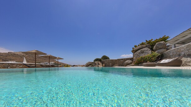 Une piscine d’un bleu éclatant, bordée de chaises longues et d’ombrelles, et entourée d’un paysage rocheux et de constructions en pierre.