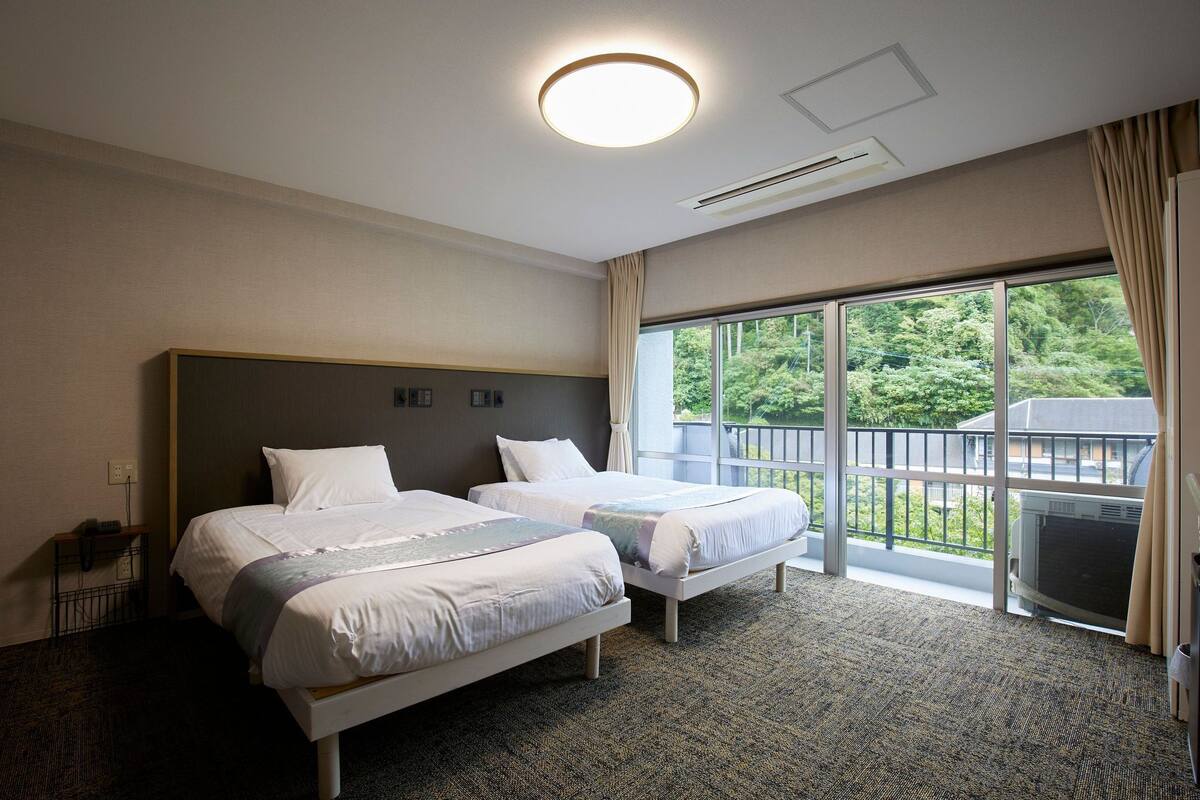 A hotel room with two beds, a large window with a view of greenery, a ceiling light, and an air conditioning unit.