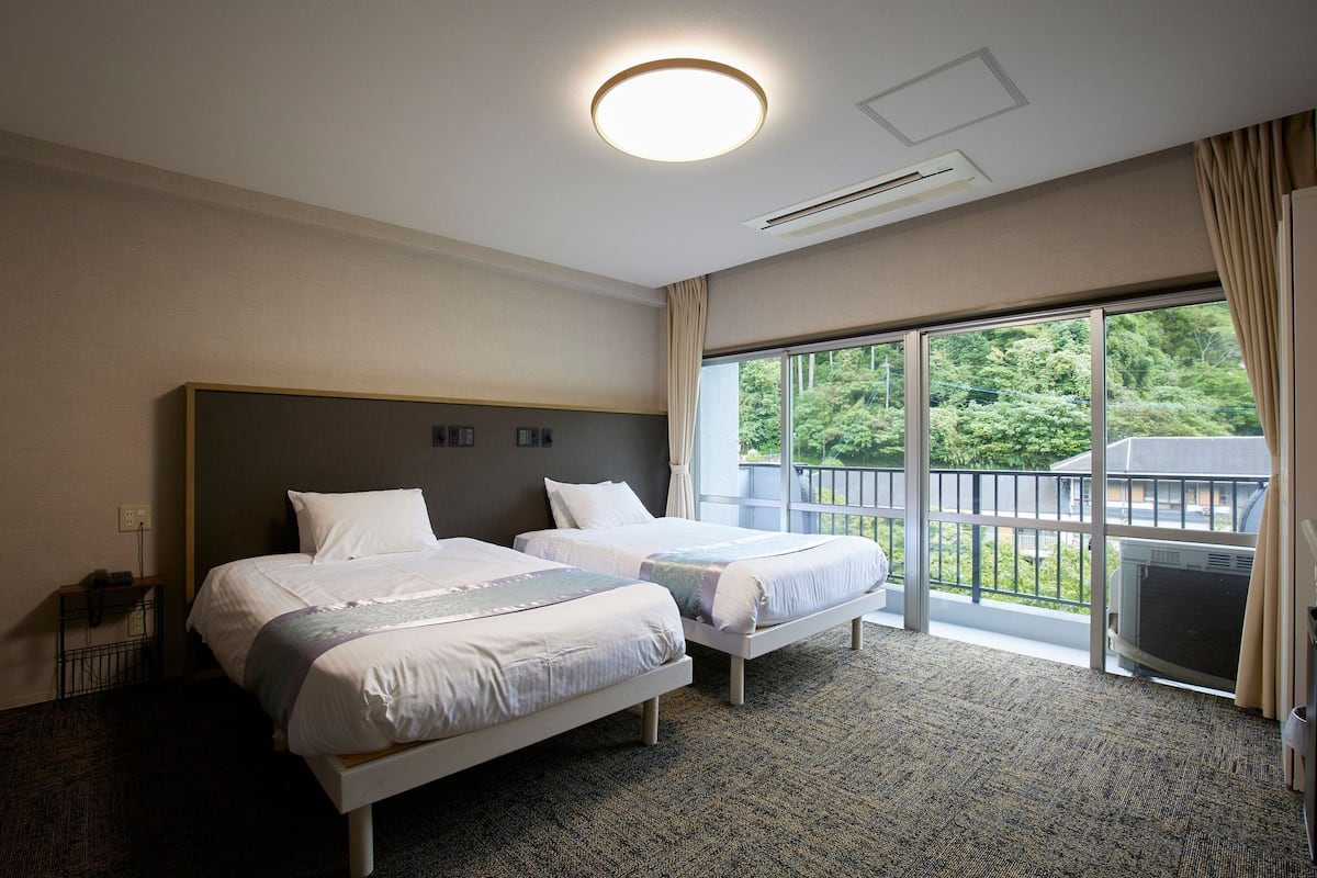 Een hotelkamer met twee bedden, een groot raam met uitzicht op groen, een plafondlamp en een airconditioning.