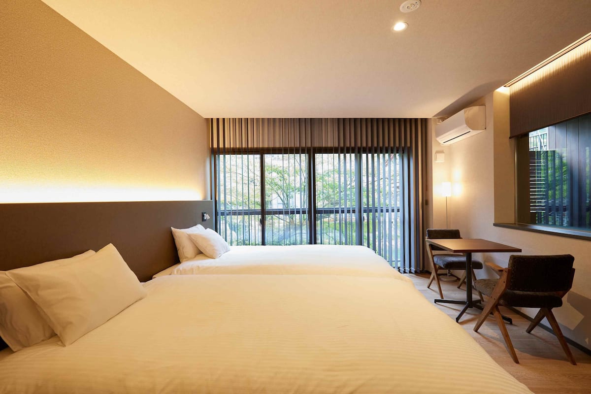 A hotel room with a bed, a table, two chairs, a window with a view, and an air conditioning unit.