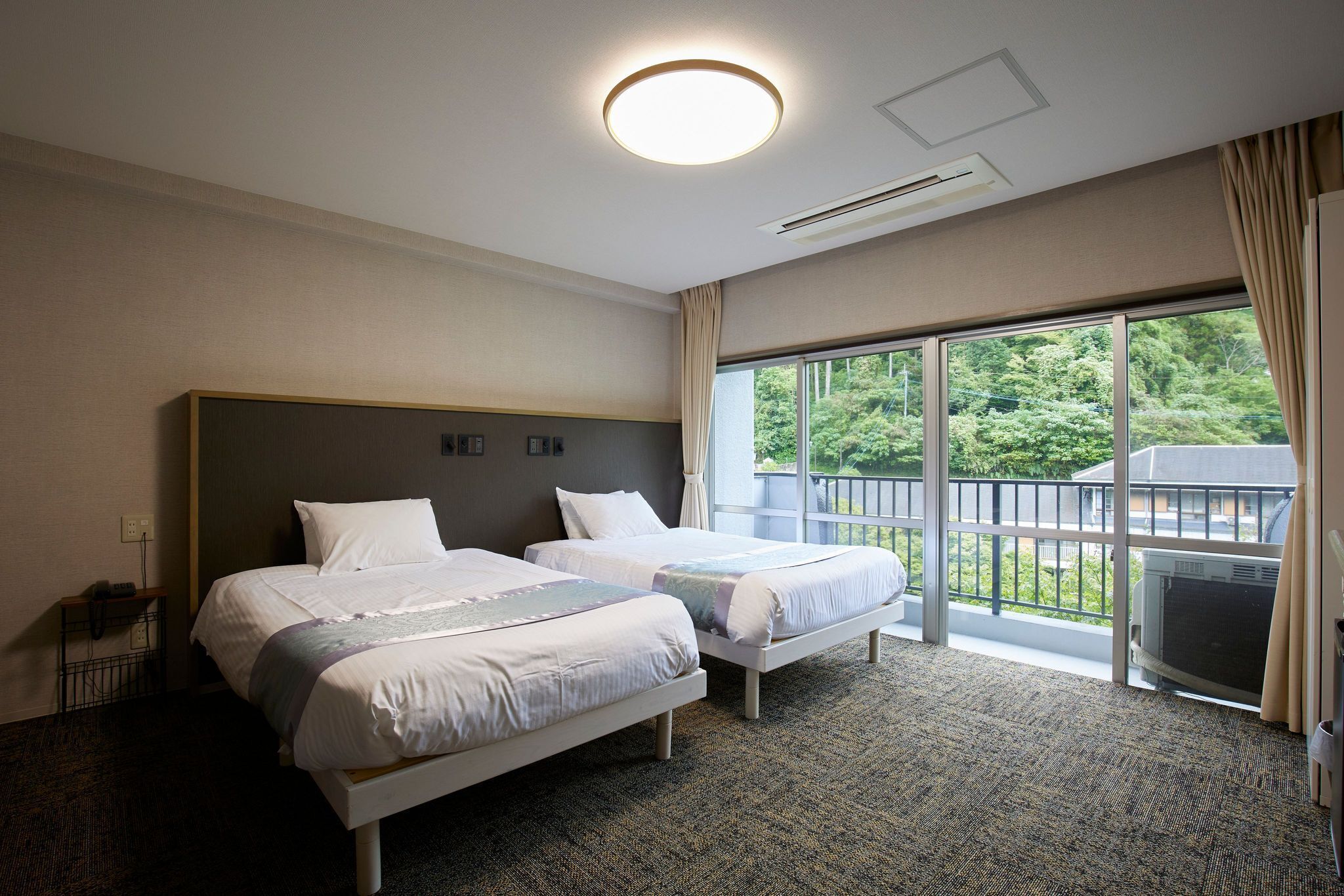 A hotel room with two beds, a large window with a view of greenery, and a ceiling light.
