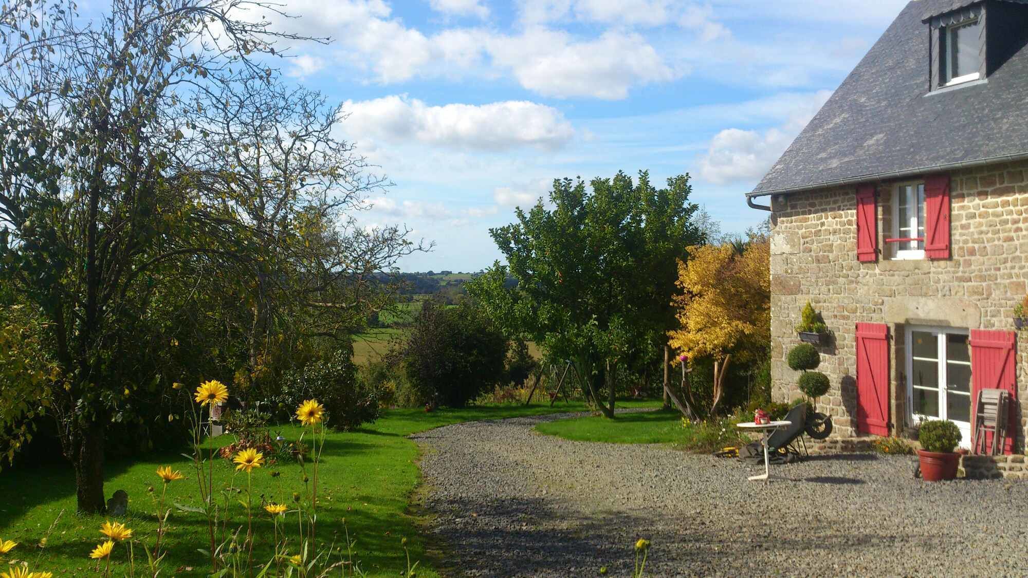 A stone house with red shutters, a gravel driveway, and a garden with yellow flowers.
