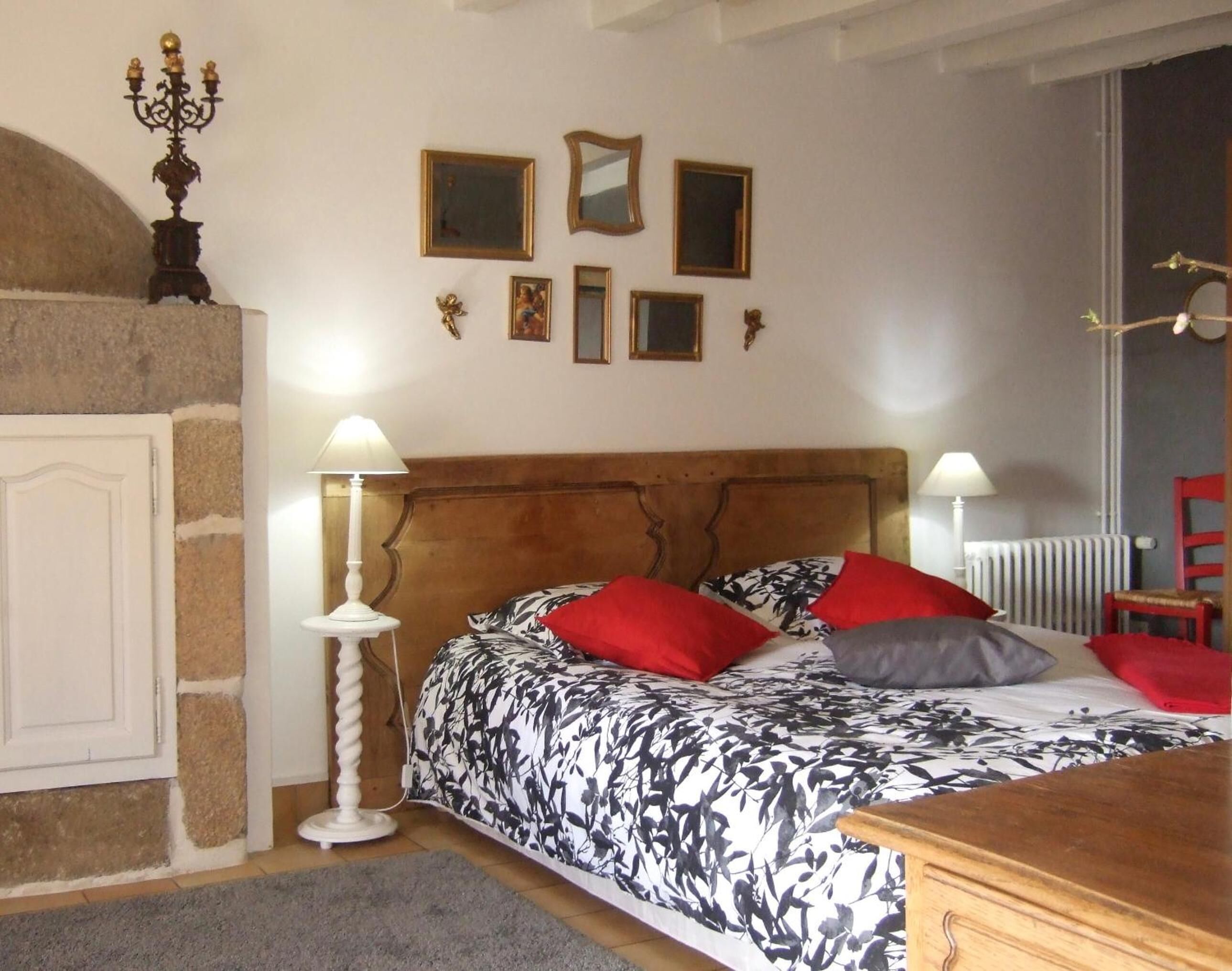 Een slaapkamer met een houten hoofdbord, een bed met bloemenbeddengoed, rode en grijze kussens, en een witte lamp naast het bed.