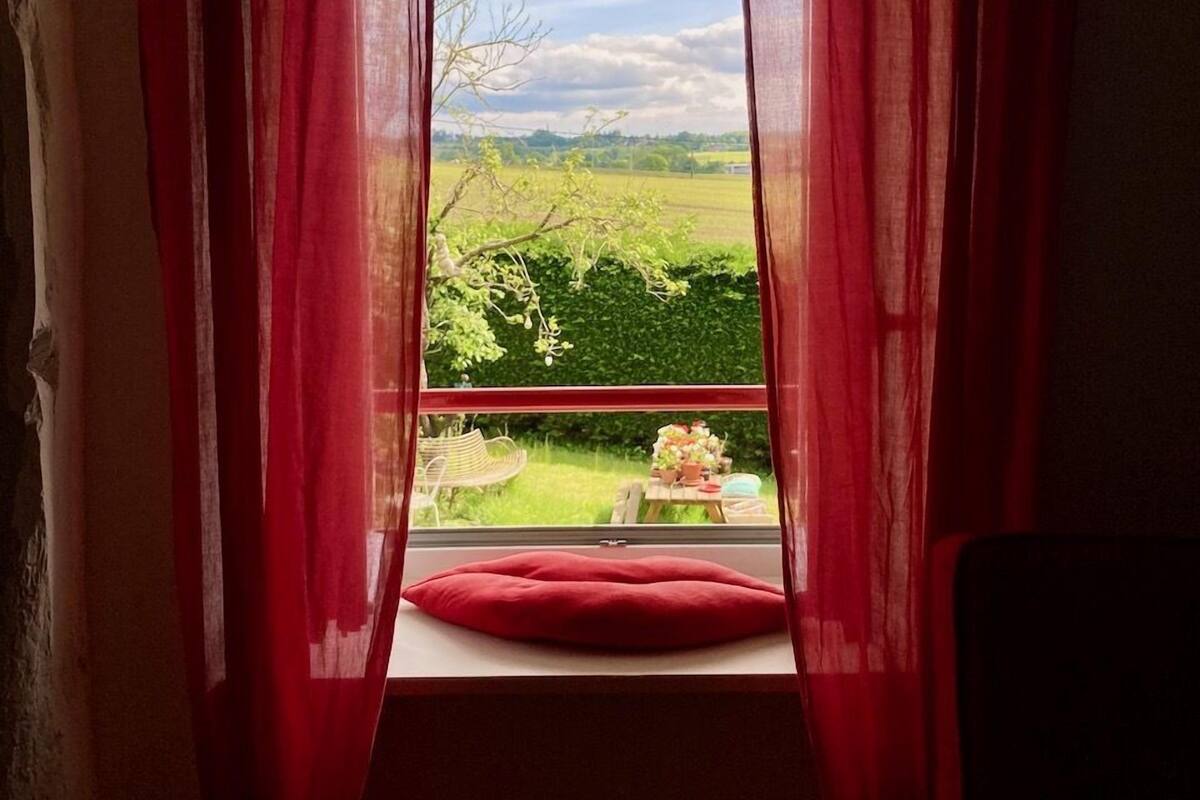 A window with red curtains, a view of a green landscape, and a red cushion on the windowsill.