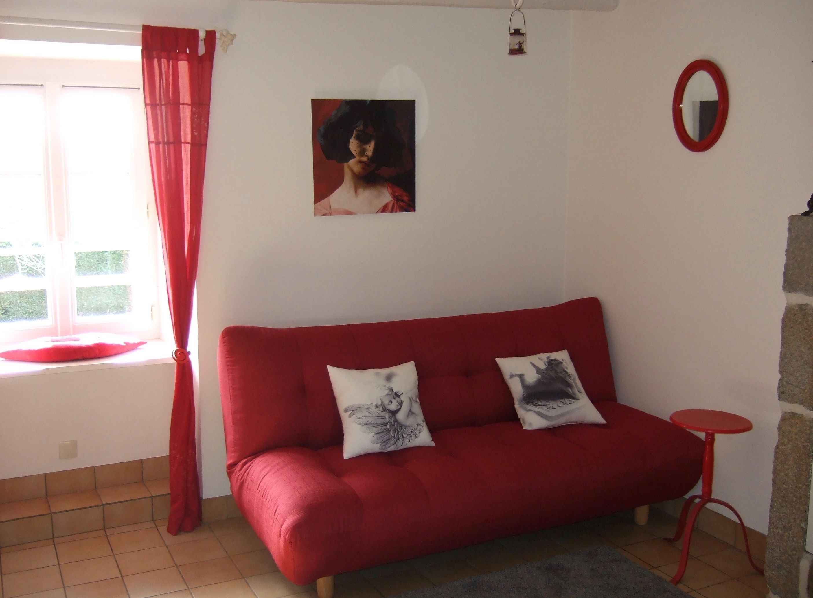 A red sofa with two decorative pillows, a small round table, a wall-mounted mirror, and a window with red curtains.