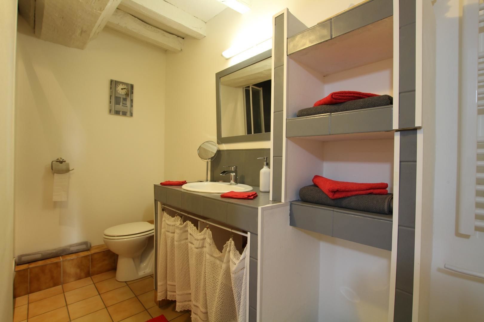 Une petite salle de bain comprenant des toilettes, un lavabo et un miroir au-dessus. On y trouve des étagères avec des serviettes et une petite horloge accrochée au mur.