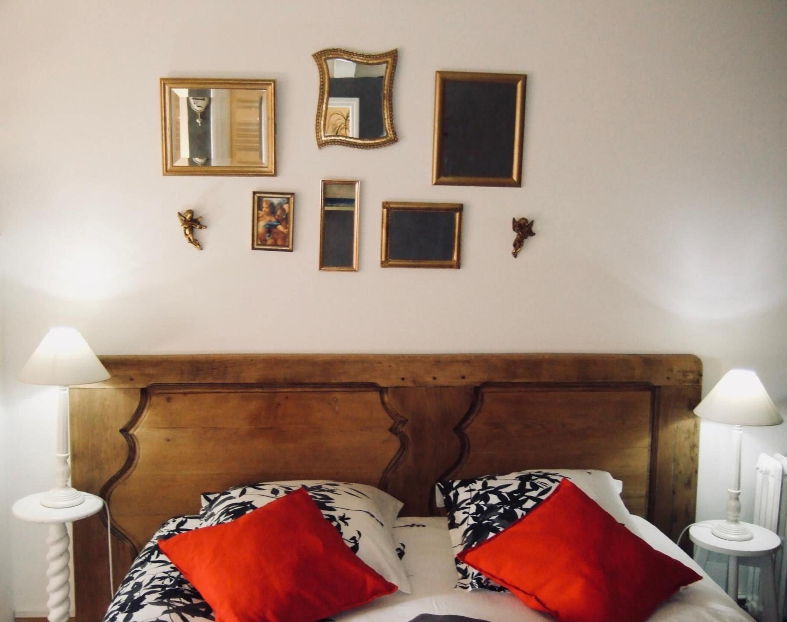 A bedroom with a wooden headboard, two bedside tables with lamps, a bed with patterned bedding and red pillows, and several framed pictures on the wall.