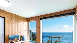 A room with wooden paneling, a white door, a framed picture, a small table with a chair, and a large window with a view of the ocean.