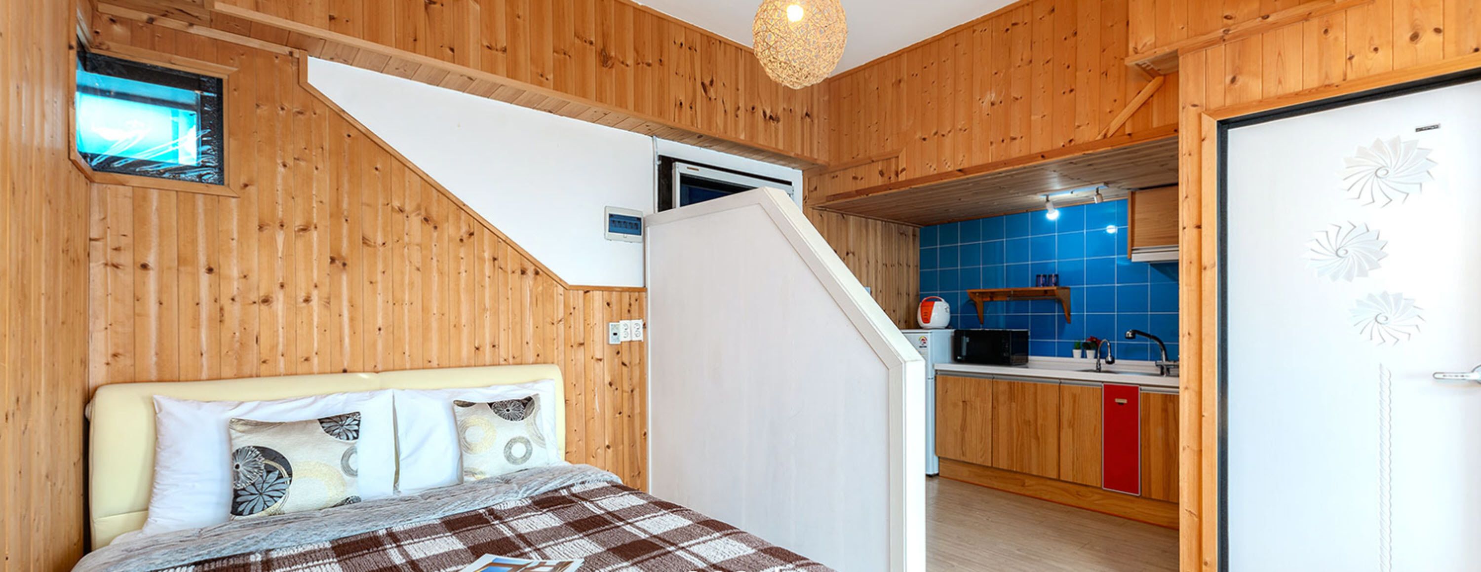 A bedroom with a bed, pillows, and a view of a kitchen with blue tiles and wooden cabinets.