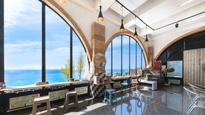 A spacious room with large arched windows overlooking the sea, a wooden bench, a small stool, and a bar area with stools and a TV.