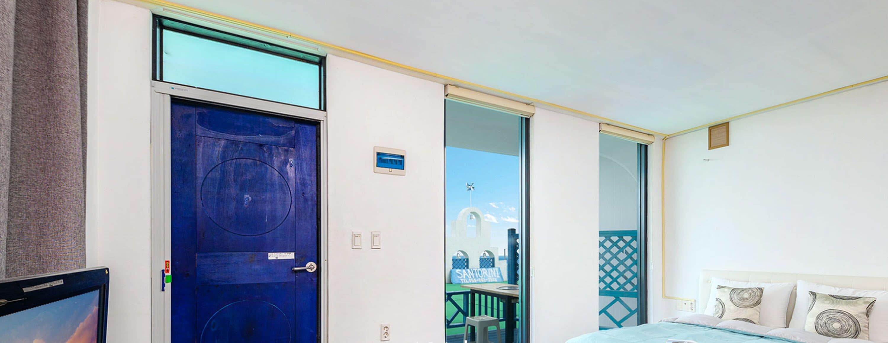 A hotel room with a large blue door, a bed with white bedding, and a view of a building through the door.