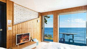 A bedroom with a large TV, a bed, and a view of the sea through a sliding door.