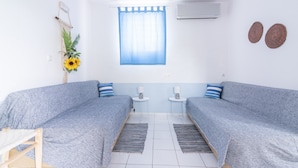Een kamer met twee grijze banken, een kleine tafel met een lamp, een raam met blauwe gordijnen en een luchtverfrisser aan de muur.