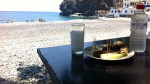 Een tafel met een bord eten, een glas water en een flesje water op een strand met een rotsachtige klif en gebouwen op de achtergrond.