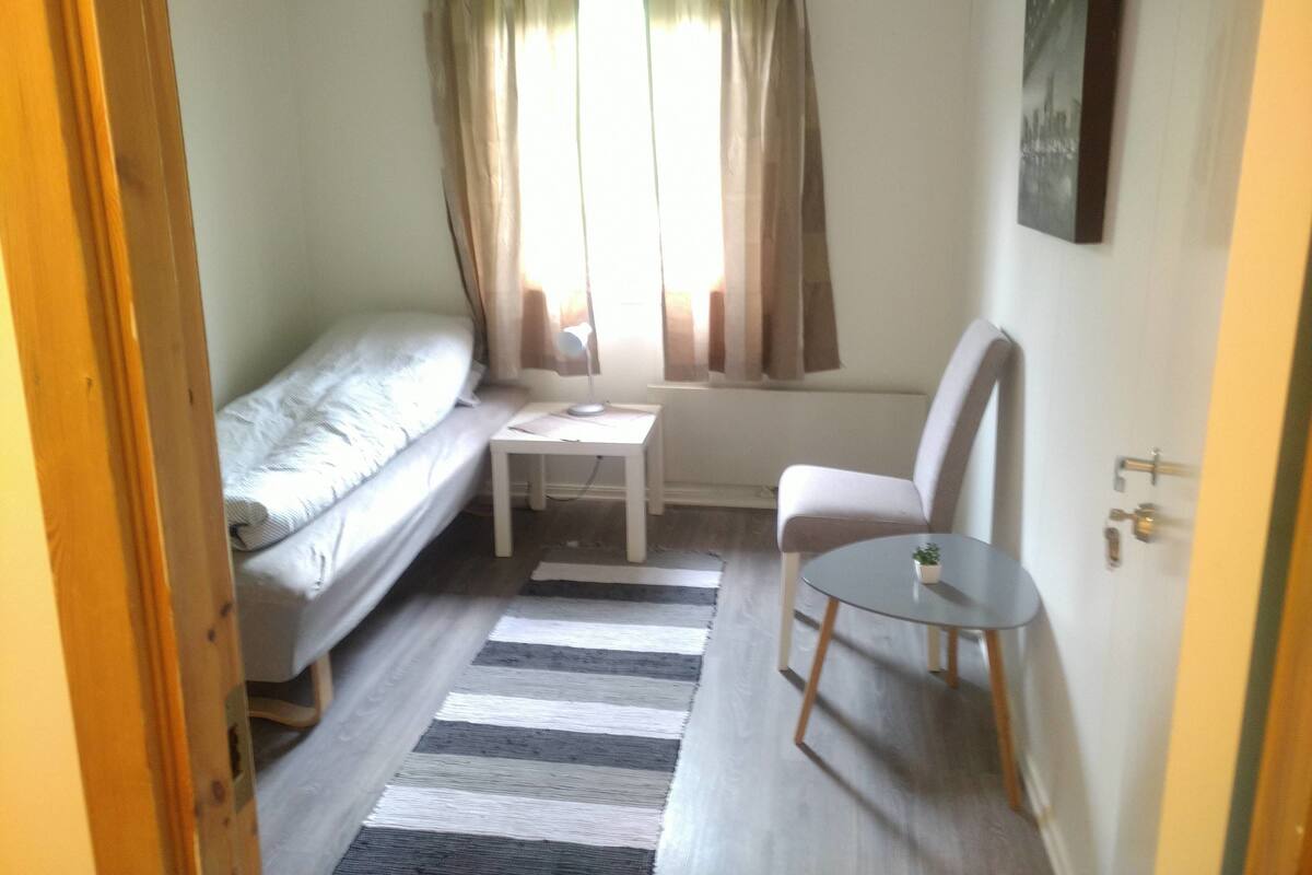 A small, compact room with a bed, a chair, a small table, and a window with curtains.