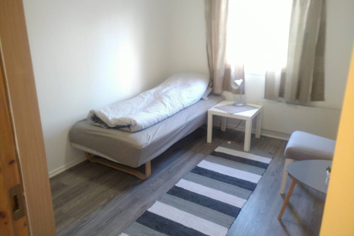 A small, neatly arranged bedroom with a single bed, a bedside table with a lamp, a striped rug, and a chair.