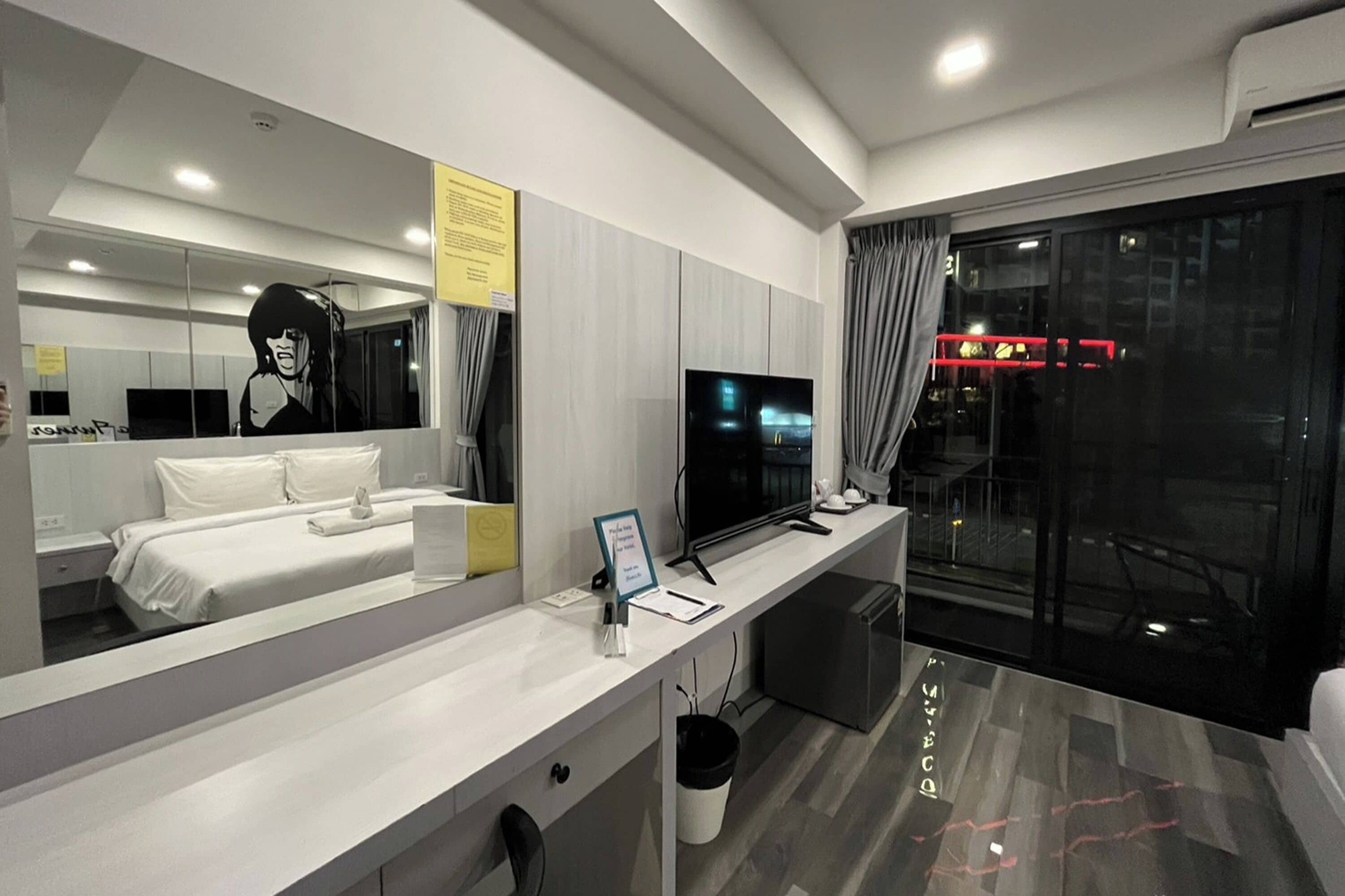 Habitación de hotel moderna con un espejo grande, una cama, un escritorio con una computadora y vistas a la ciudad a través de un ventanal amplio.