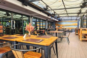 Een eethoek met houten tafels en stoelen, grote ramen en uitzicht op zee.