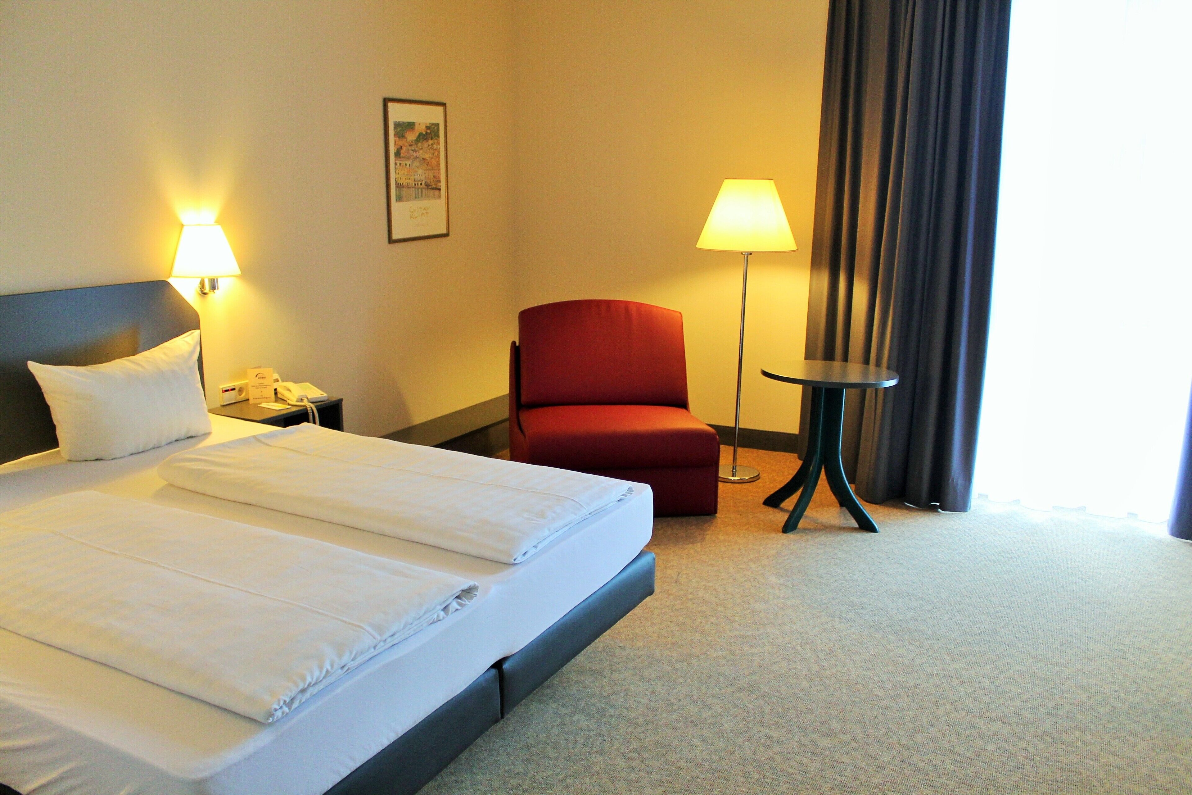 A hotel room with a bed, a red chair, a small table, and two wall lamps.