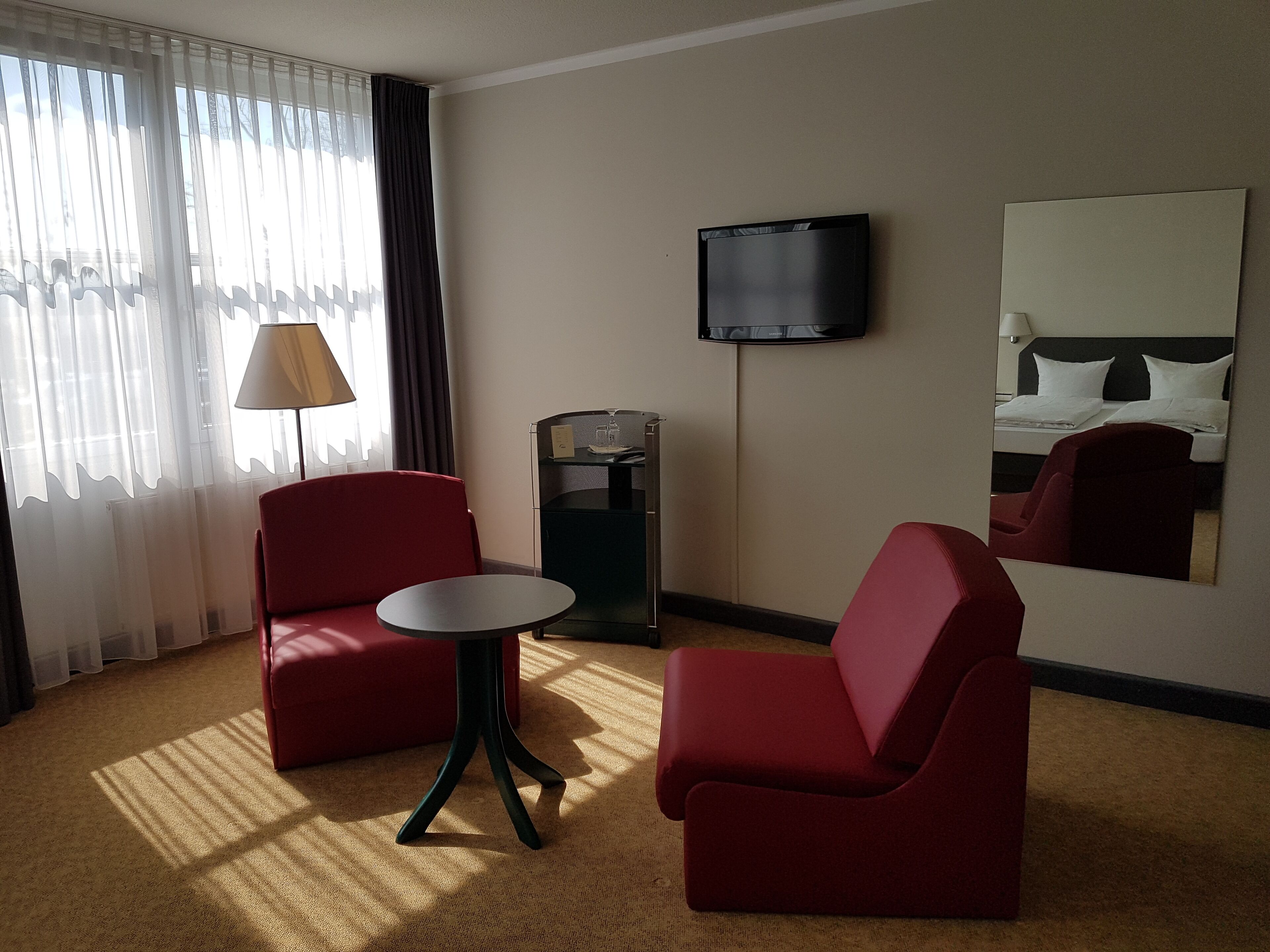 Een hotelkamer met twee rode fauteuils, een kleine ronde tafel, een aan de muur bevestigde televisie en een grote spiegel die het bed weerspiegelt.