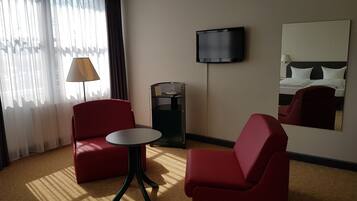 Een hotelkamer met twee rode fauteuils, een kleine ronde tafel, een aan de muur bevestigde televisie en een grote spiegel die het bed weerspiegelt.