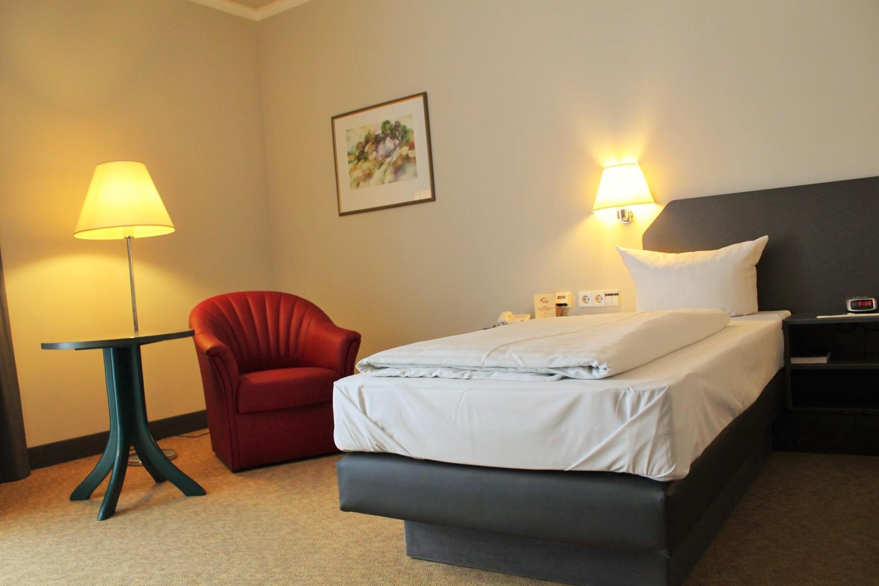 A hotel room with a bed, a red chair, a table with a lamp, and wall-mounted lights.
