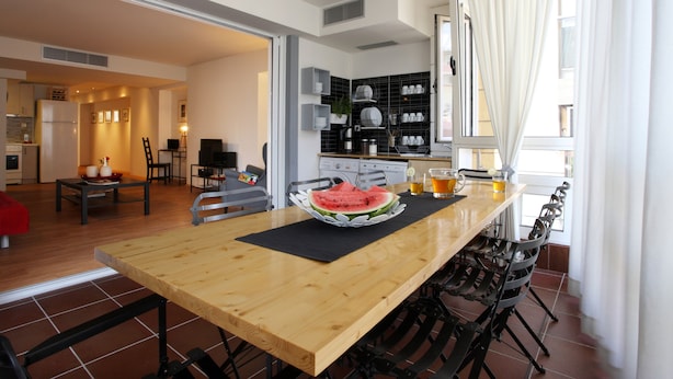 Une cuisine moderne avec une table à manger en bois, des chaises noires et un tapis noir. Il y a une pastèque sur la table et deux verres de jus d’orange.