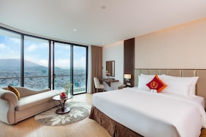 A hotel room with a large bed, a sitting area, a desk, and a view of a cityscape through large windows.