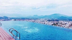 An infinity pool with a wooden deck, overlooking a cityscape with mountains in the background. - Regalia Gold (Nha Trang)