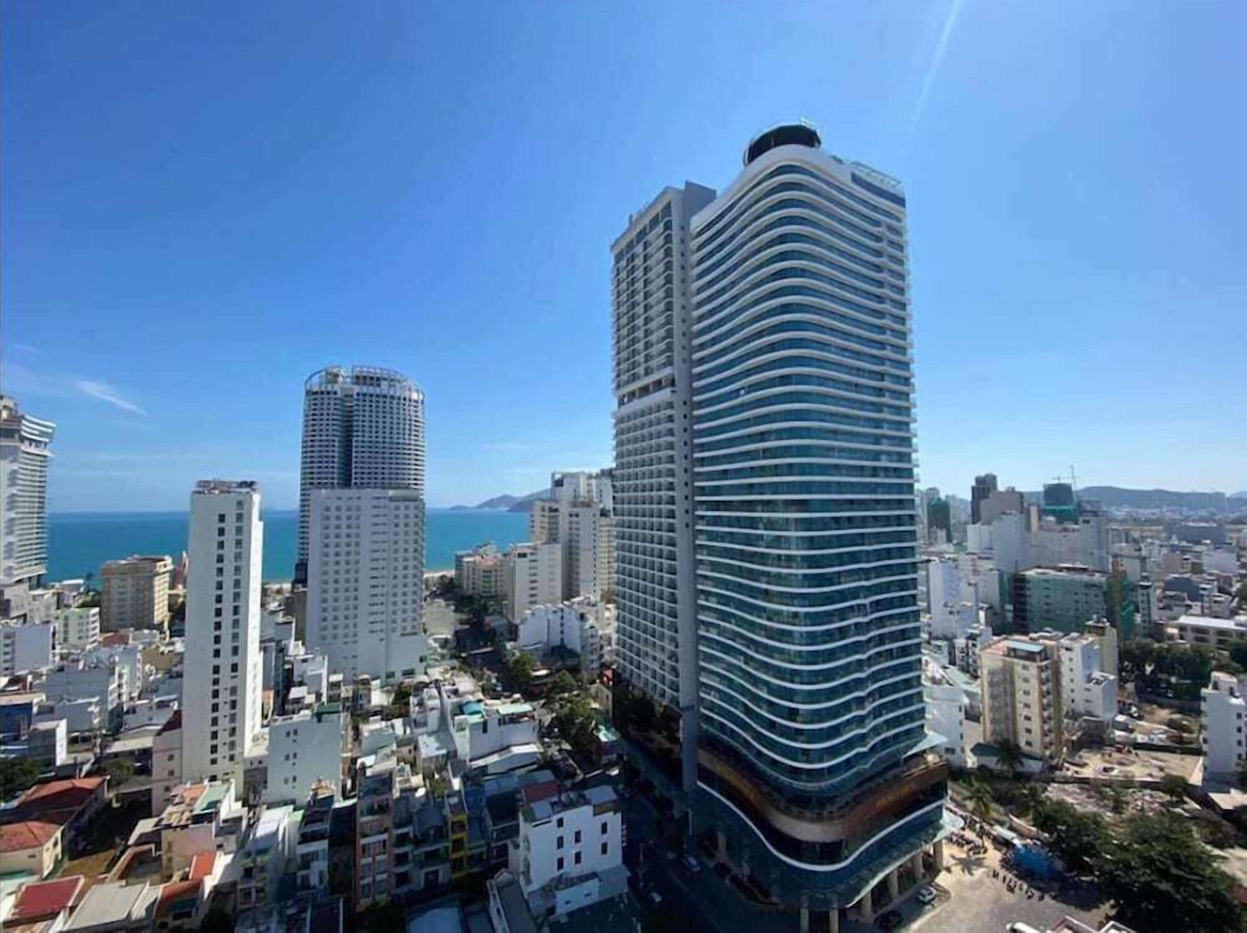 A high-rise building with a curved facade, surrounded by other buildings and a clear view of the sea.