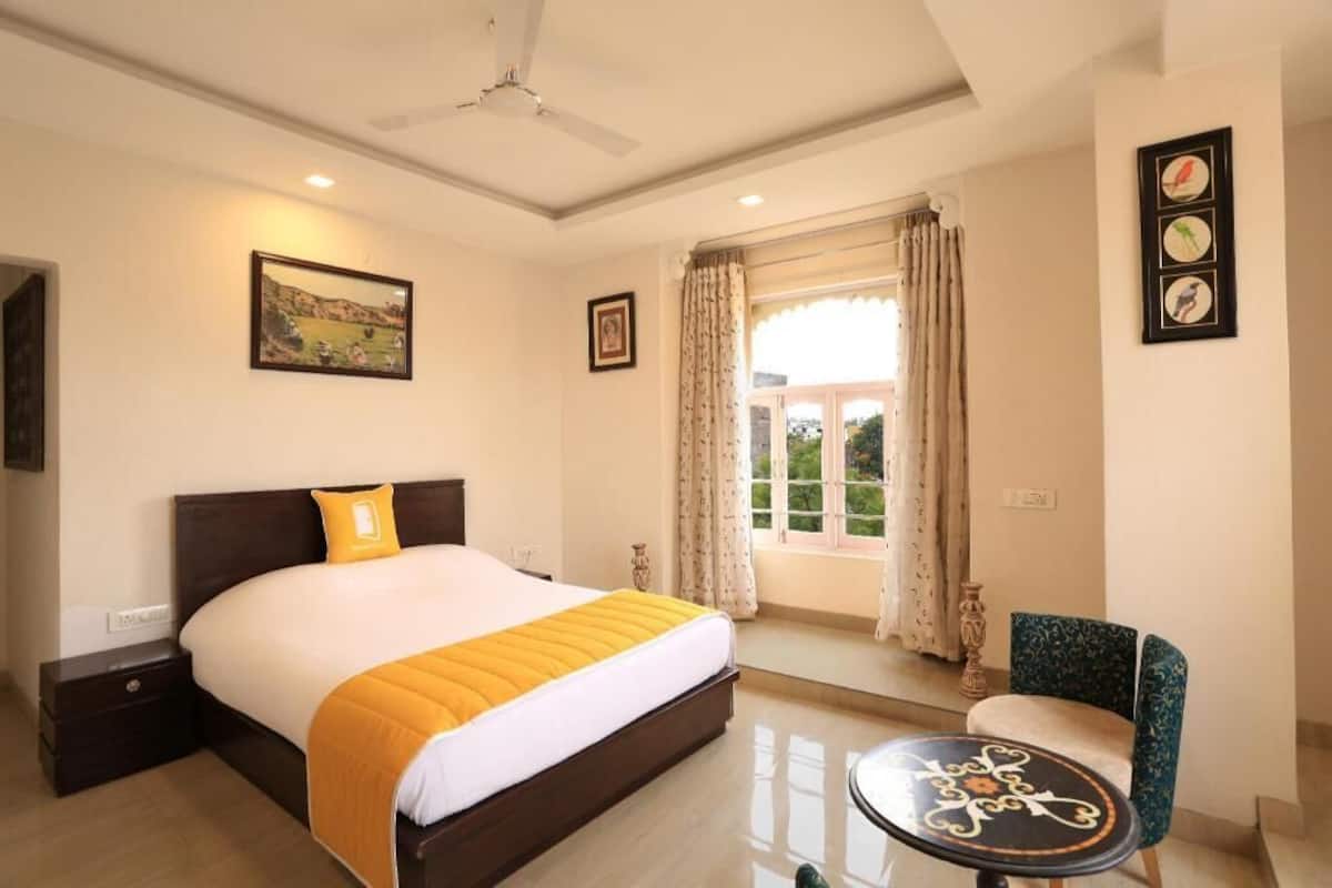 A hotel room with a bed, a chair, a table, a window with curtains, and a ceiling fan.