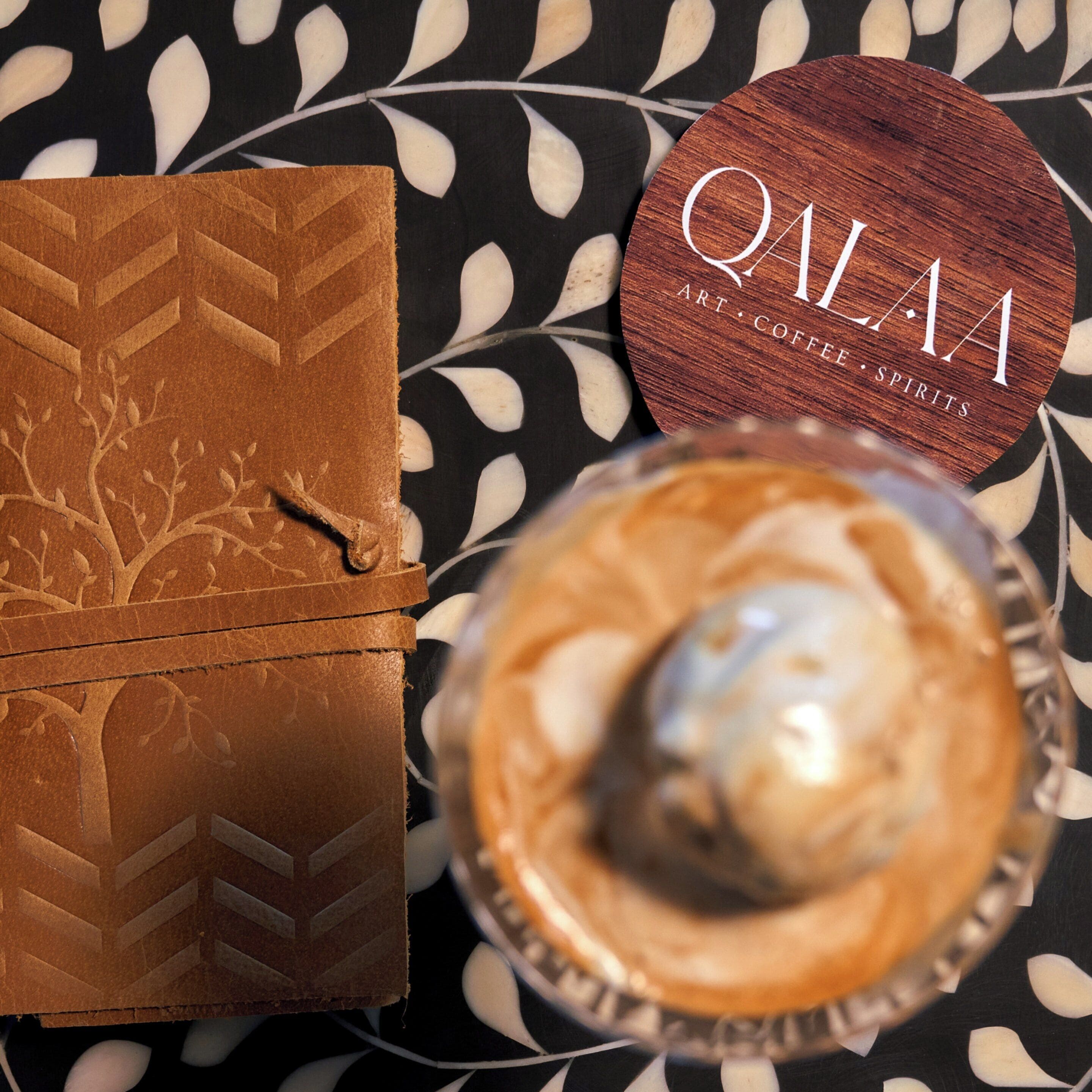 A wooden box with a tree design, a round wooden coaster with "QWAM ART COFFEE SPIRITS" text, and a glass of coffee with a spoon.